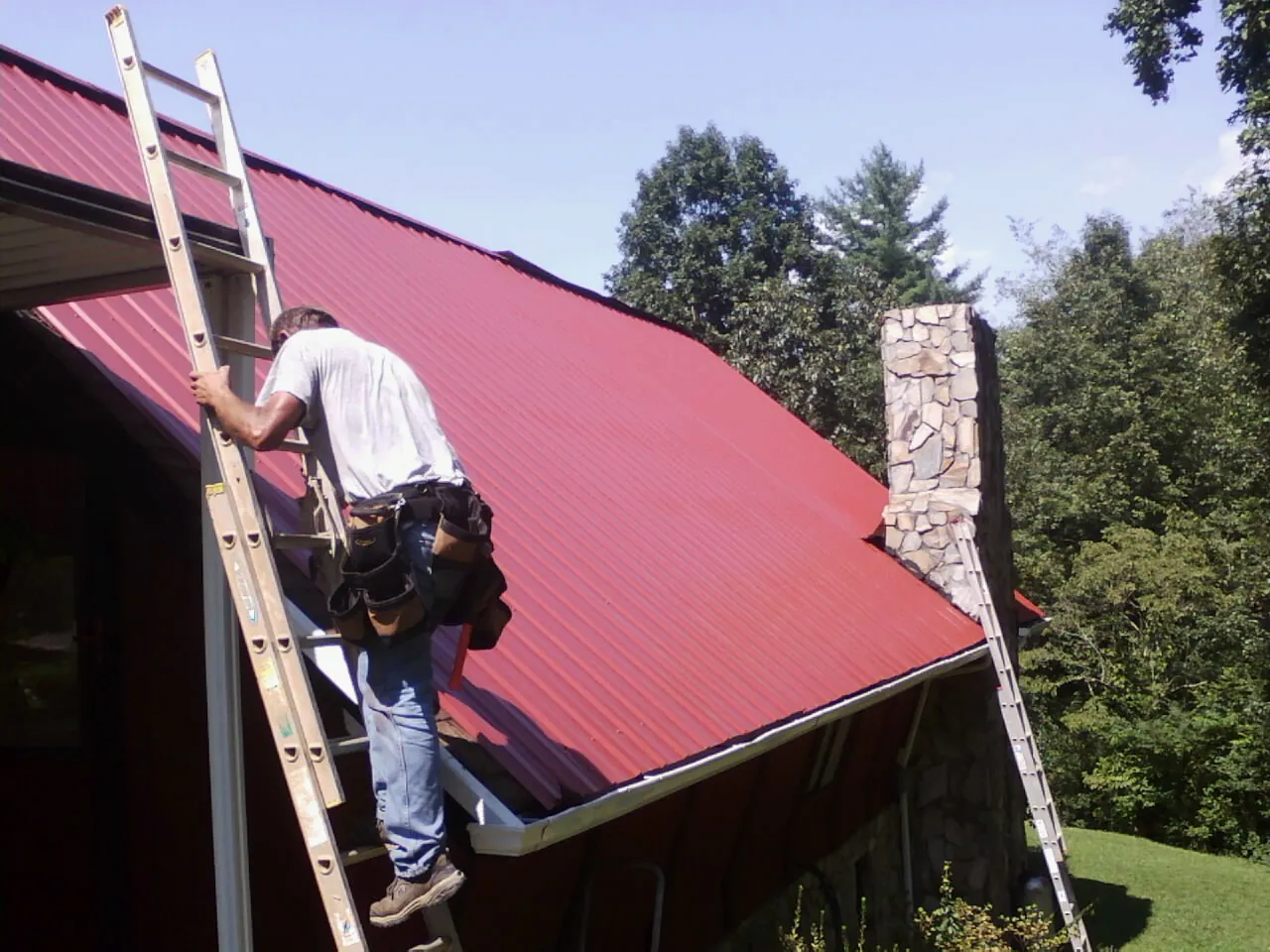 Roofer on a ladder beside a red metal roof and stone chimney — Soffit & Fascia Repair by Roofing Brothers in Valley Springs